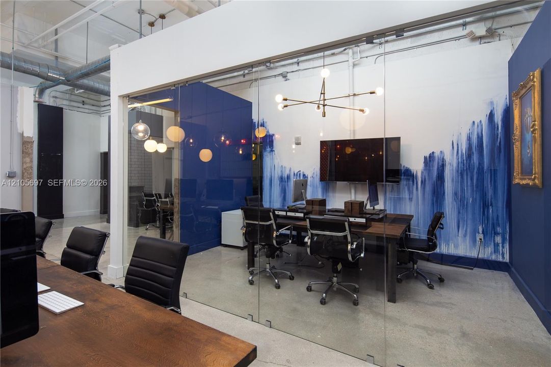 Enclosed office space featuring full-height glass partitions, polished concrete flooring, exposed industrial ceiling, and a sculptural multi-arm chandelier