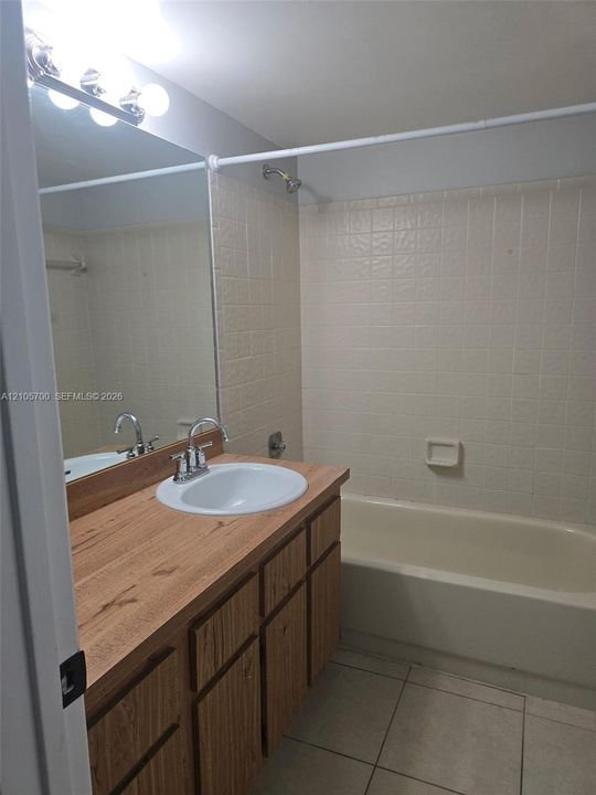 Bathroom featuring a wood-finish vanity cabinet, a solid-surface countertop with an integrated basin, a full-width vanity mirror, tile flooring, and a tiled shower surround