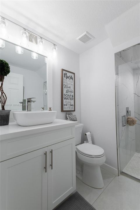 White vanity featuring a rectangular vessel sink with chrome faucet, topped with a solid surface counter and accompanied by a walk-in shower with floor-to-ceiling tile surround and a glass door