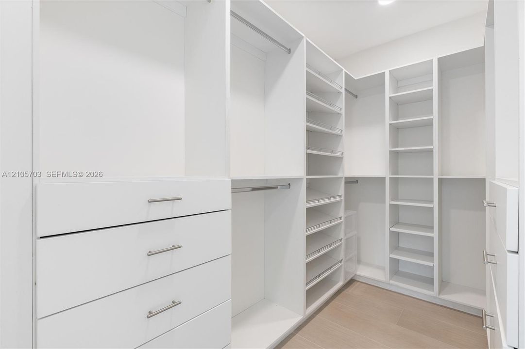 Luxurious walk-in closet featuring custom white cabinetry with integrated drawers, extensive shelving throughout, polished chrome hardware, and warm wood-finish flooring