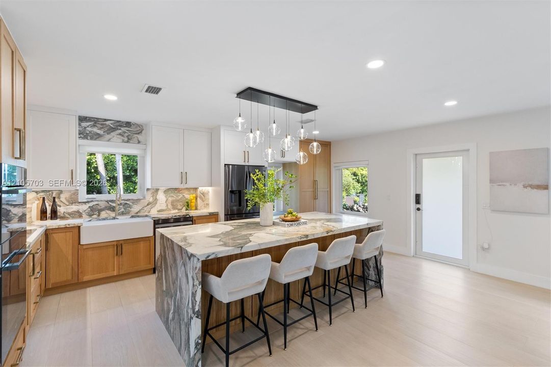 Kitchen featuring a waterfall-edge island with stone surfacing, apron-front farmhouse sink, custom cabinetry with dual-tone finish, built-in stainless steel appliances, and multi-pendant lighting fixture