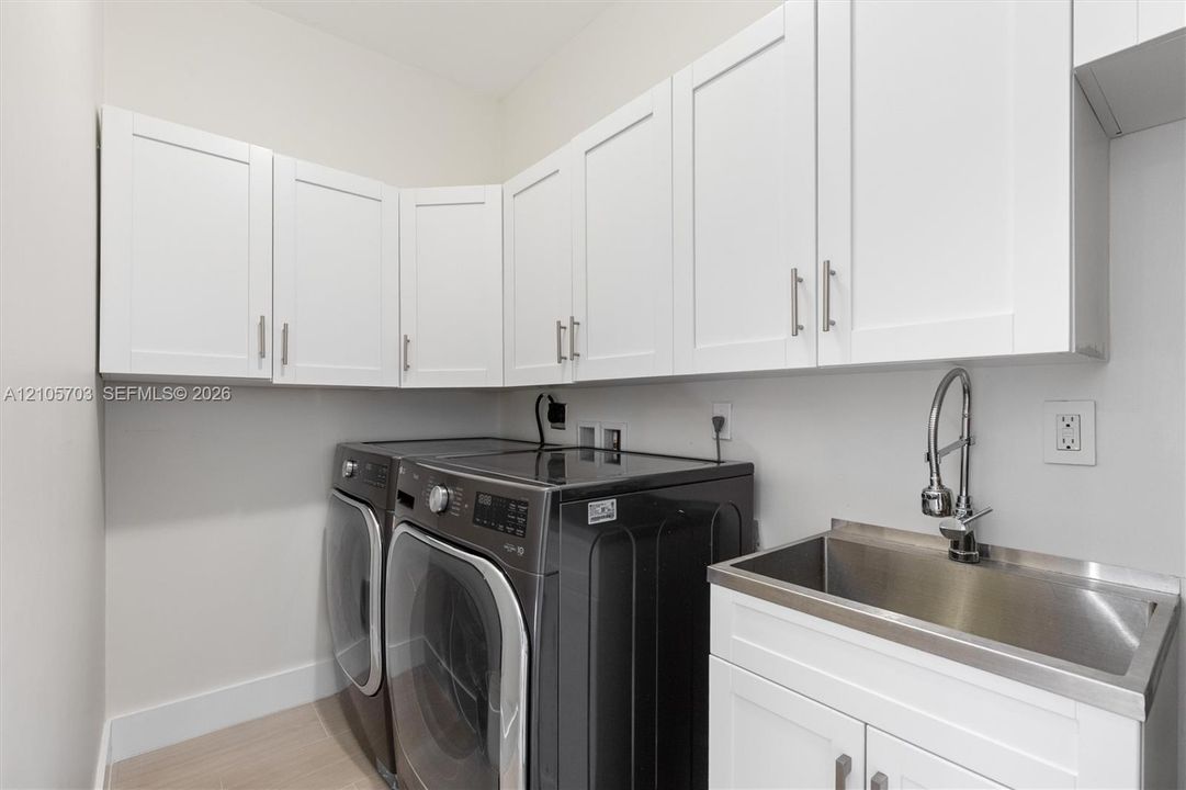 Laundry room featuring white shaker-style wall cabinetry, stainless steel utility sink with a high-arch pull-down faucet, and light-toned flooring