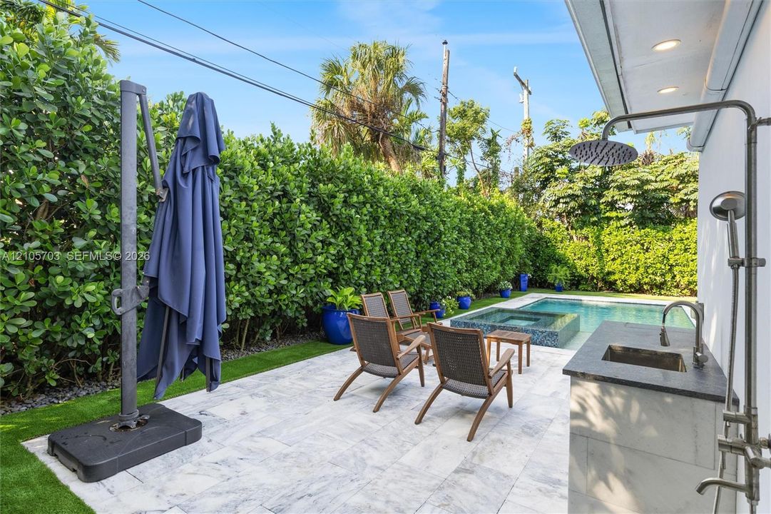Outdoor patio featuring natural stone paving, an integrated wet bar with a dark stone countertop, a wall-mounted shower fixture, an in-ground swimming pool with an elevated spa, and mature privacy hedging