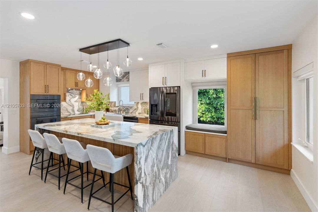 Kitchen featuring a large waterfall island with patterned stone surfacing, natural wood-finish cabinetry, built-in dual ovens, multi-light pendant fixture, and a window-side built-in bench
