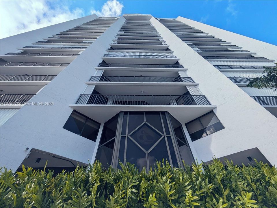High-rise building exterior featuring a white concrete façade, tiered private balconies with metal railings, floor-to-ceiling glass paneling, and a geometric-patterned entryway