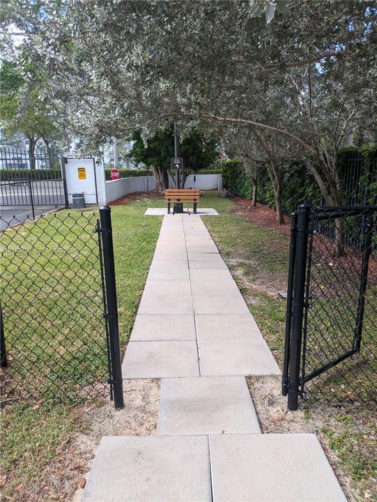 Fenced outdoor space featuring a paved walkway, mature trees, and a permanent wooden bench