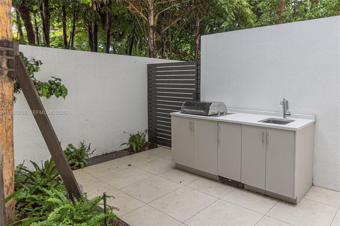 Outdoor kitchen area featuring large-format tile flooring, a stone-finish countertop, stainless steel integrated grill, undermount sink, and privacy wall