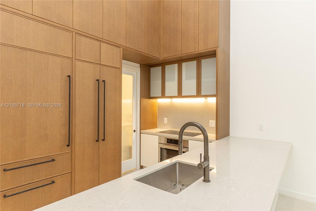 Floor-to-ceiling wood-finish cabinetry, integrated refrigerator, stone countertop with waterfall edge, undermount stainless steel sink, and glass-paneled upper cabinets