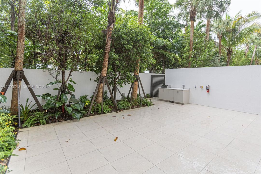 Private outdoor patio featuring light-toned tile flooring, built-in cabinetry with a sink, stainless steel grill, solid privacy walls, and mature perimeter landscaping