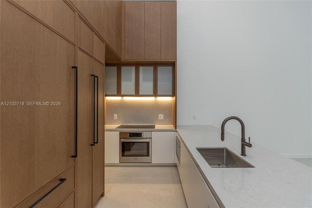 Kitchen featuring full-height wood-finish cabinetry, white solid surface countertops, a stainless steel wall oven, an integrated cooktop, and an undermount sink with a high-arch pull-down faucet