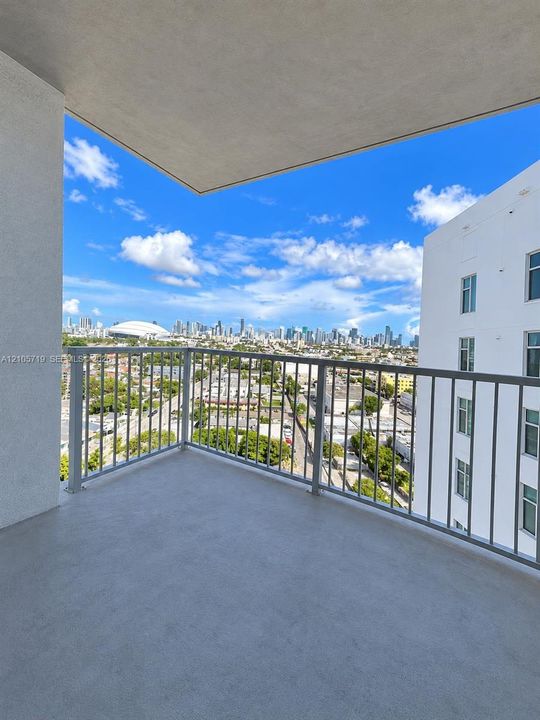 Covered balcony featuring concrete flooring and a metal railing, overlooking an urban skyline