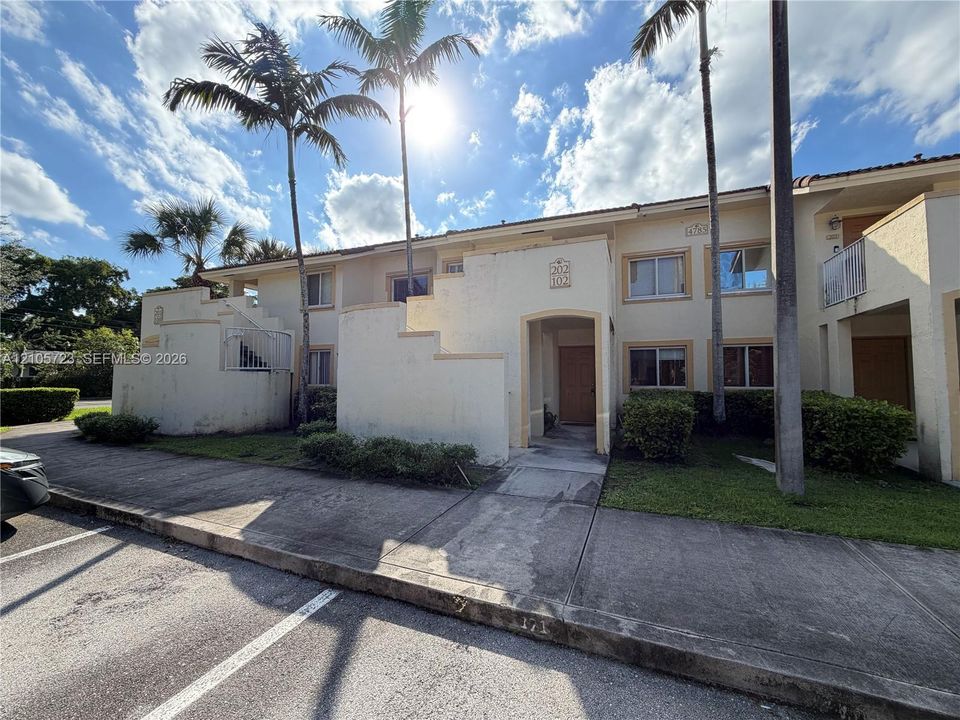 Two-story exterior featuring a light-colored stucco facade, recessed entryways, metal balcony railings, and manicured landscaping with established palm trees