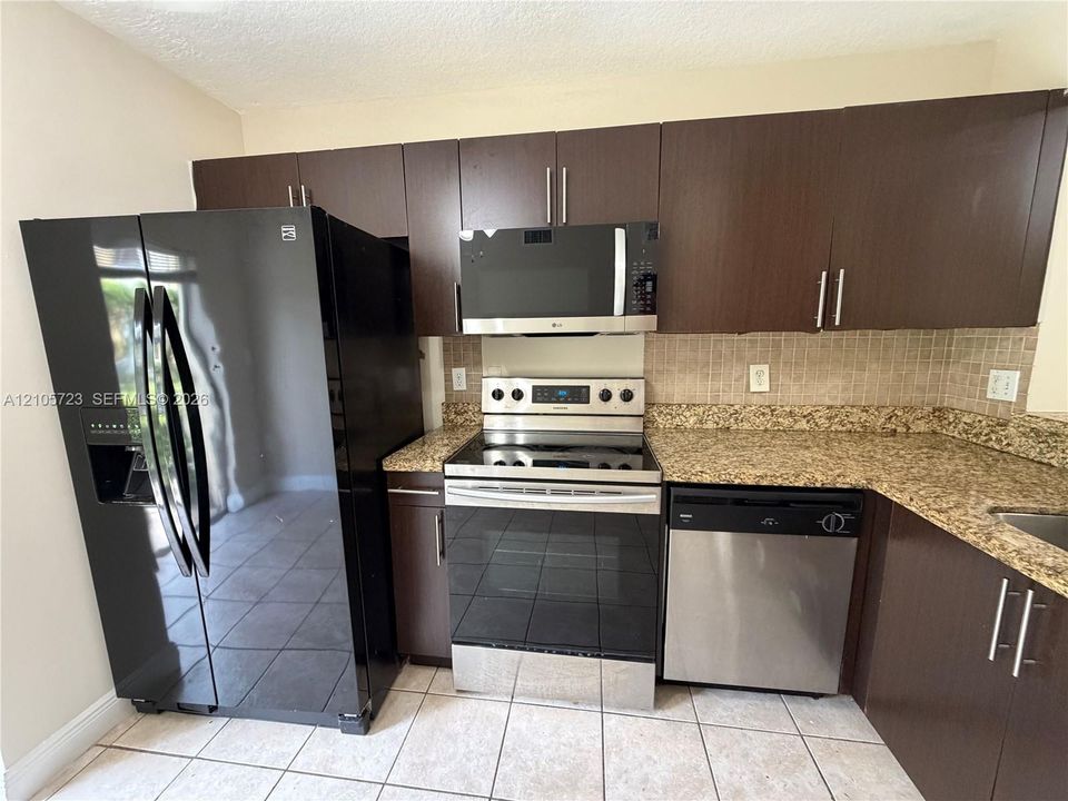 Kitchen featuring dark-finish cabinetry, stone-style countertops, mosaic tile backsplash, and a suite of stainless steel and black appliances, including a built-in microwave and dishwasher