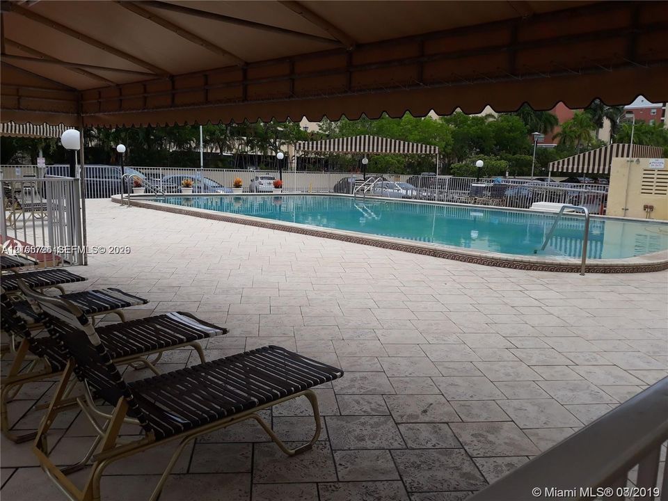 Swimming pool with paved surround, featuring a shaded canopy area and metal perimeter fencing