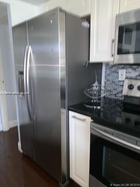 Stainless steel side-by-side refrigerator, white shaker-style cabinetry, dark solid-surface countertop, glass mosaic tile backsplash, and stainless steel built-in microwave