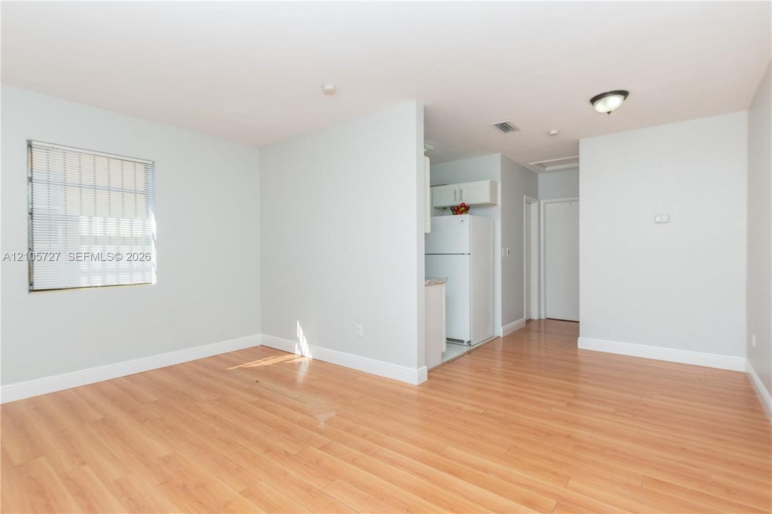 Living area featuring wood-finish flooring, neutral wall tones, and recessed lighting, with a partial partition wall separating the space from the kitchen area containing white cabinetry and a refrigerator