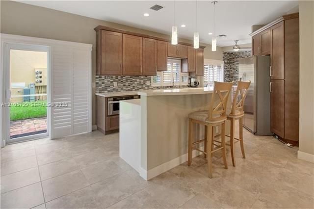 Kitchen featuring wood cabinetry, quartz-topped peninsula, multi-colored stone tile backsplash, large-format tile flooring, and sliding glass door with plantation shutters