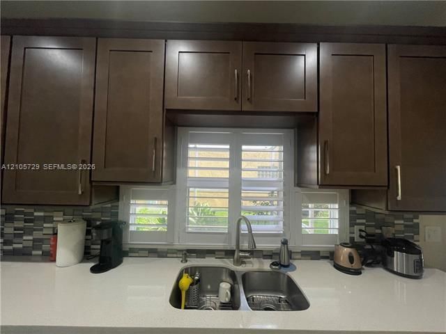Dark wood-finish cabinetry featuring shaker-style doors, stone-surface countertops, multi-toned tile backsplash, stainless steel double-basin undermount sink, and white plantation shutters