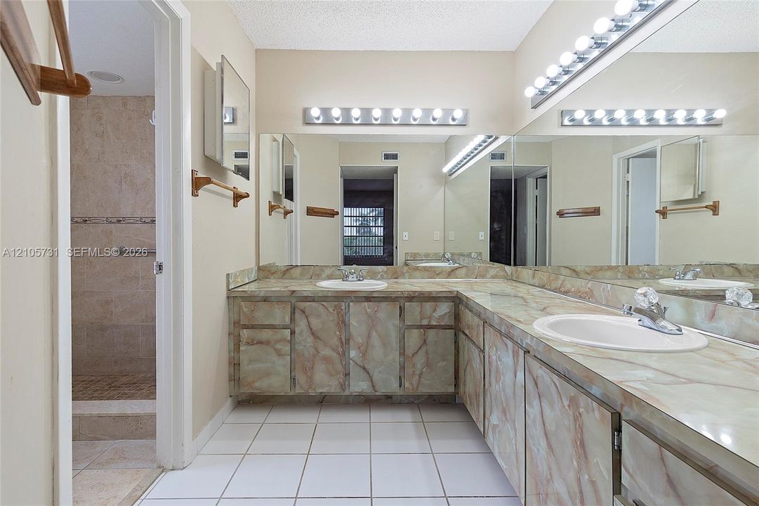 L-shaped vanity featuring custom-fitted stone-finish cabinetry and countertops, dual white drop-in sinks, extensive full-width wall mirrors, and integrated multi-bulb lighting fixtures