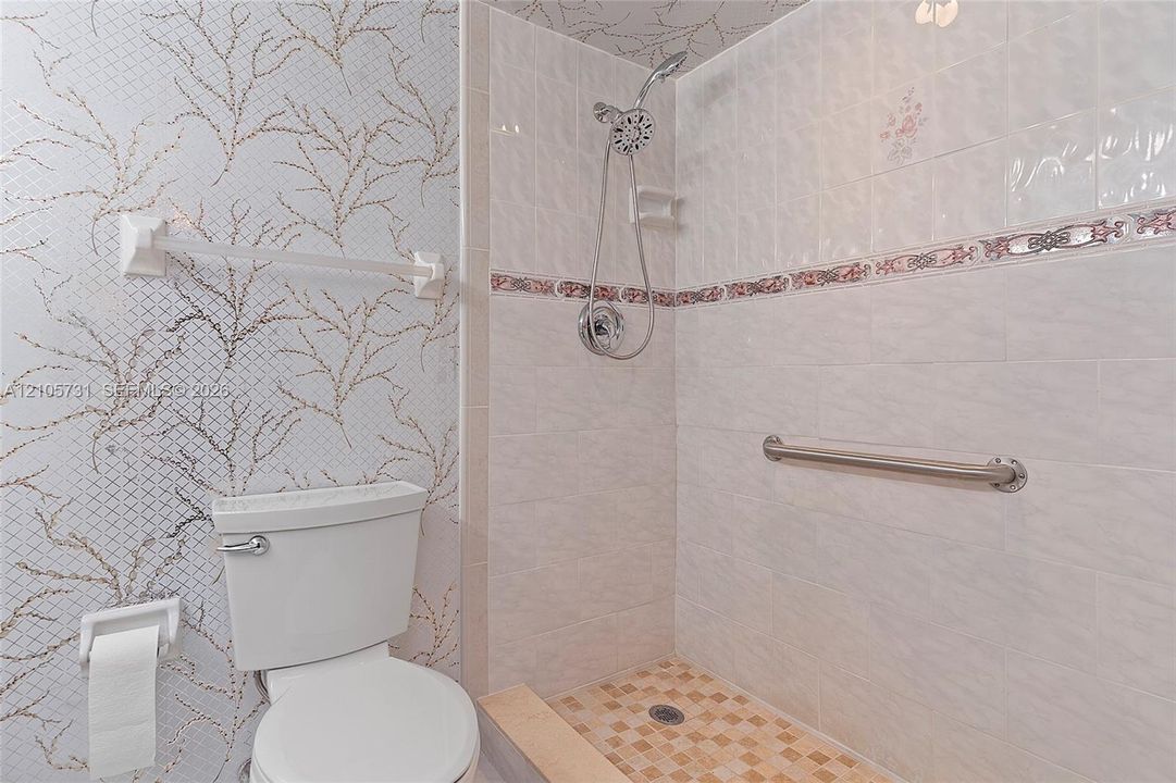 Bathroom featuring a tiled walk-in shower with chrome hand-held showerhead, integrated metallic accent tile border, chrome grab bar, and patterned wall covering