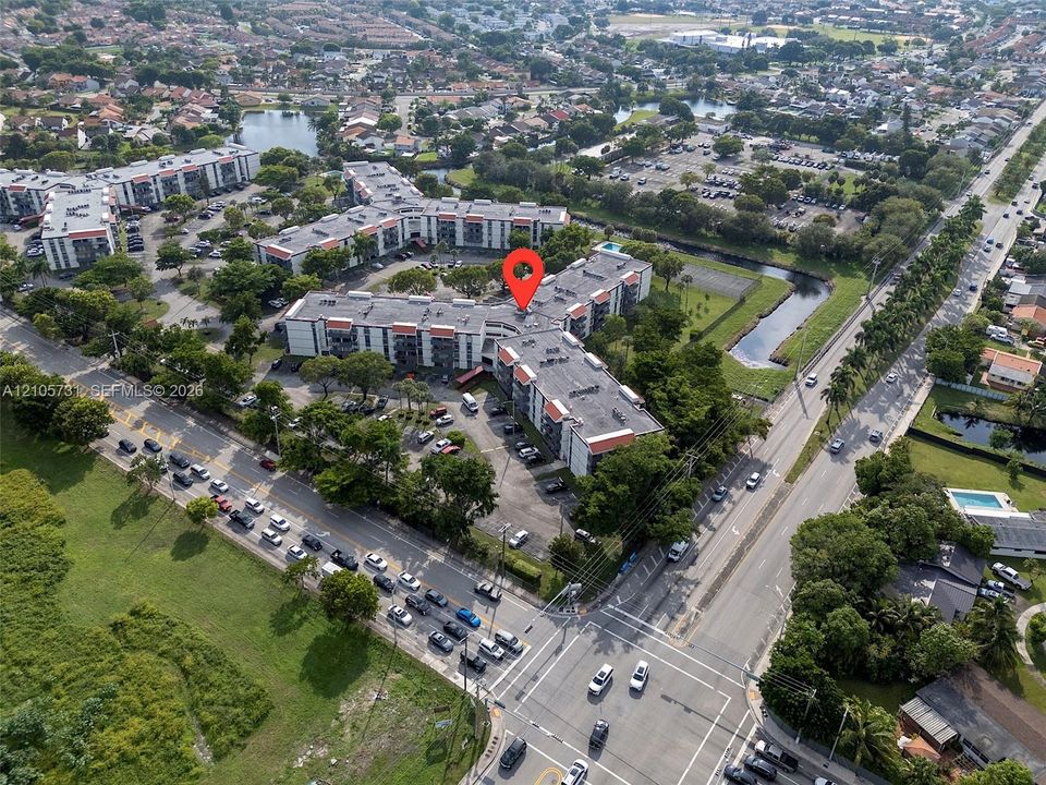 Aerial perspective of a multi-story residential building featuring asphalt roofing, exterior balconies, surface parking, and surrounding landscape areas
