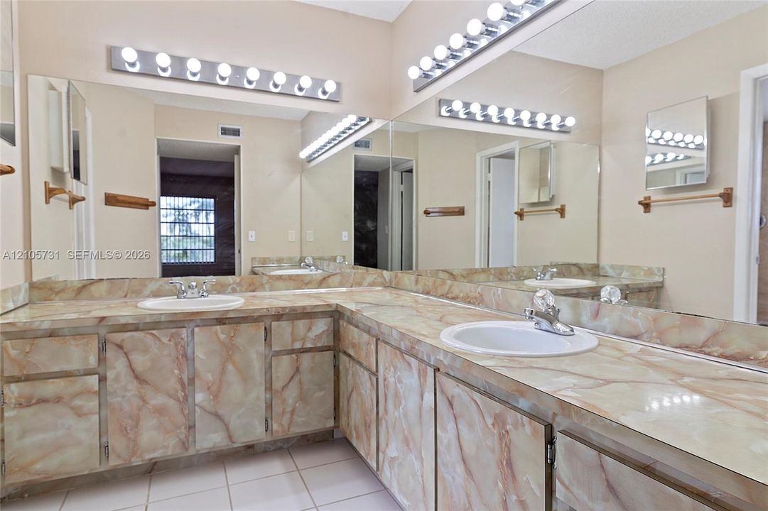 Dual-sink vanity featuring patterned stone-finish countertops, custom cabinetry with matching stone-finish doors, expansive wall-to-wall mirror, and multi-bulb theatrical lighting fixtures