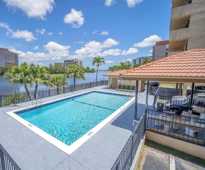 In-ground swimming pool with a concrete deck, covered pavilion featuring a tiled roof, metal perimeter fencing, and water frontage