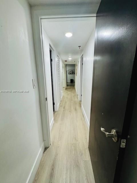 Hallway featuring wood-finish flooring, white door casings, integrated ceiling lighting, and a dark-toned interior door with a brushed metal lever handle