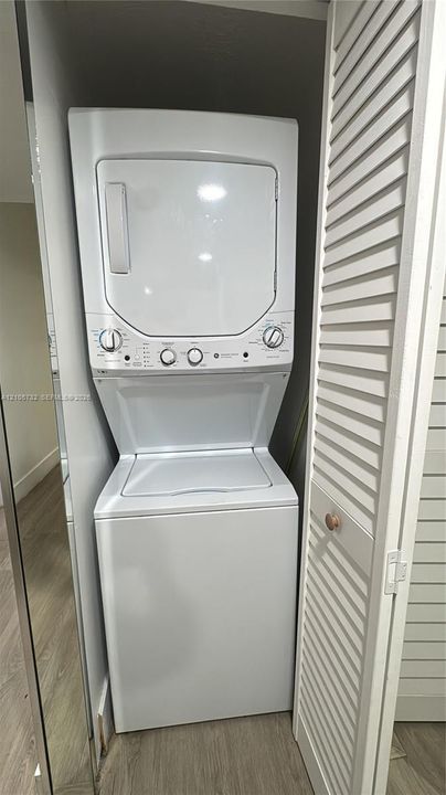 Stacked white washer and dryer unit housed within a closet featuring white louvered doors and wood-finish flooring