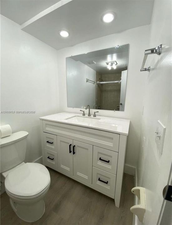 White vanity featuring shaker-style cabinetry, matte black hardware, integrated sink, and stone-surface countertop, complemented by wood-finish flooring and a wall-mounted mirror