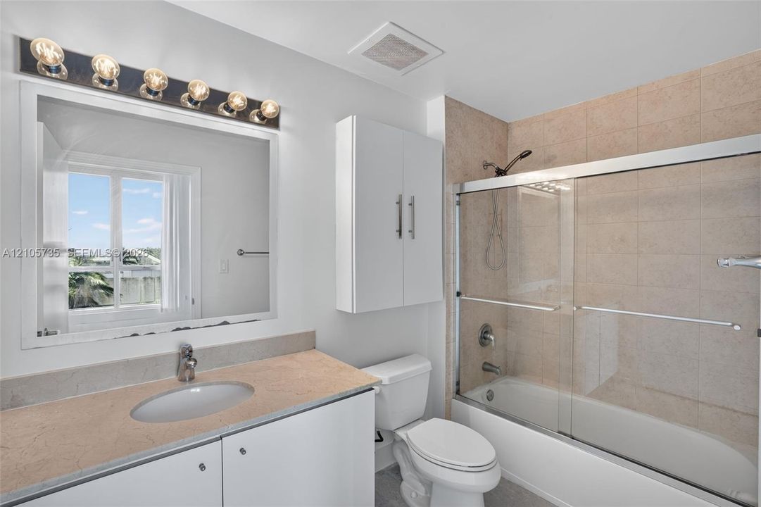 Bathroom featuring a vanity with a solid-surface countertop, an integrated oval sink, a wall-mounted storage cabinet, and a bathtub-shower combination with a glass sliding enclosure and stone-tile surround