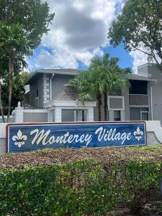 Community entrance featuring a monument sign, stucco-finished building facade, quoined corner detailing, and mature palm trees