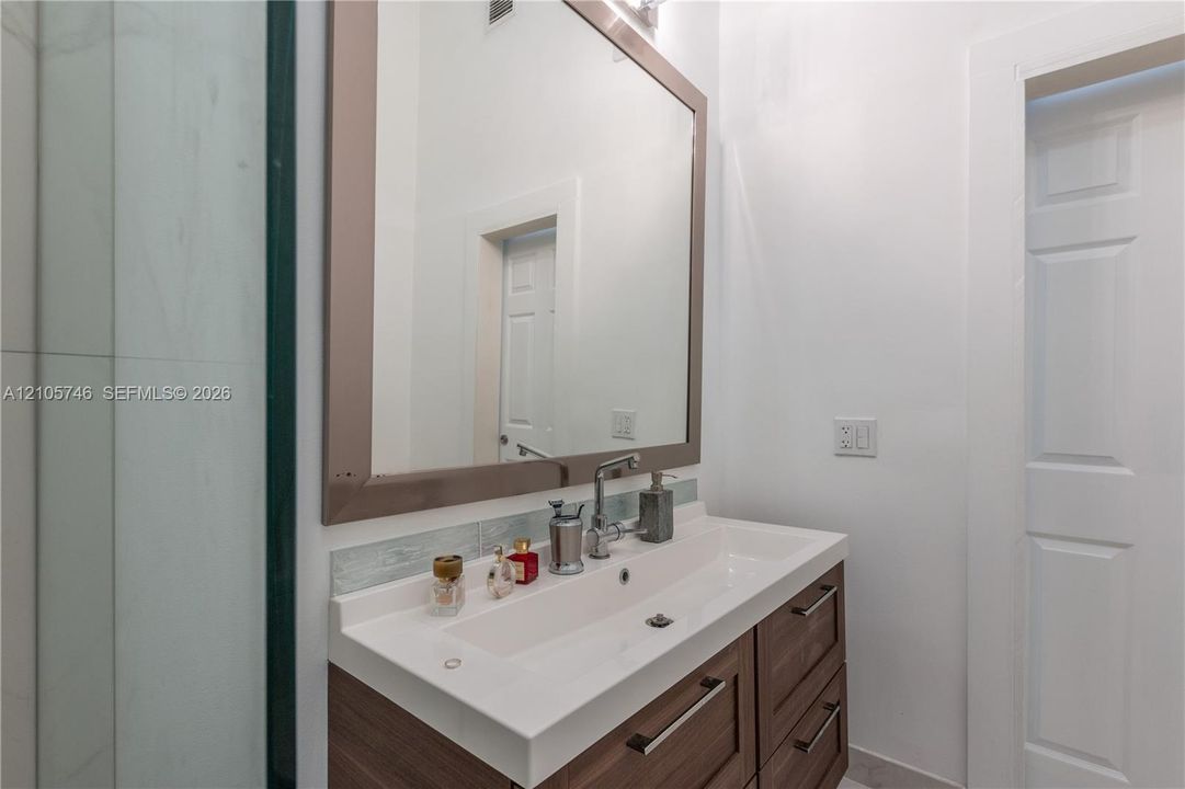 Wood-finish vanity with a rectangular sink, stone-composite countertop, full-width mirror with a wide frame, and wall-mounted tile backsplash
