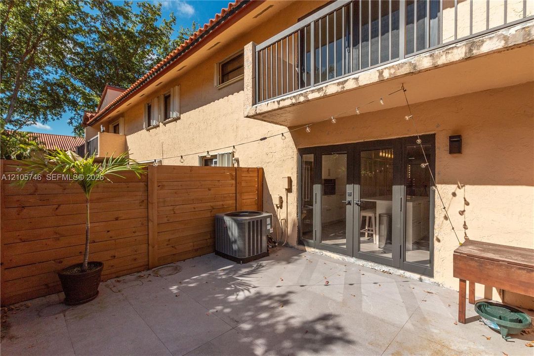 Exterior patio featuring concrete pavers, horizontal wood privacy fencing, and dark-framed glass multi-panel doors, complemented by a second-level balcony with metal railings