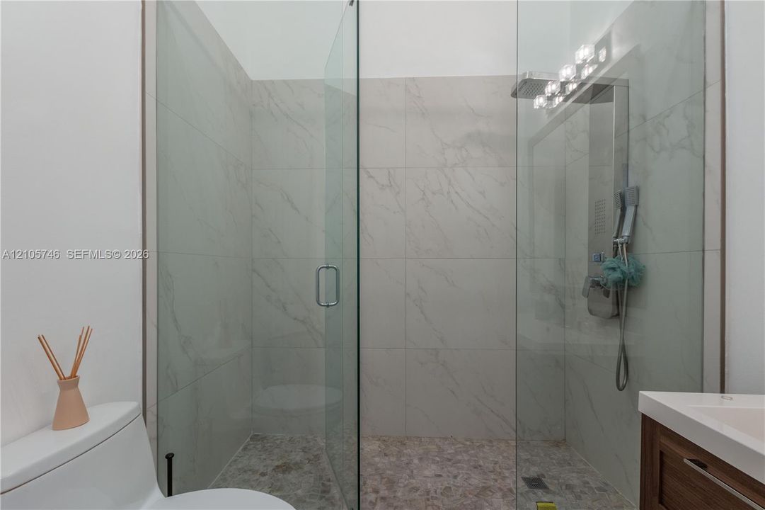Walk-in shower featuring large-format stone-tile walls, pebble-stone flooring, a frameless glass enclosure, and a chrome rainfall showerhead with integrated hand-held sprayer