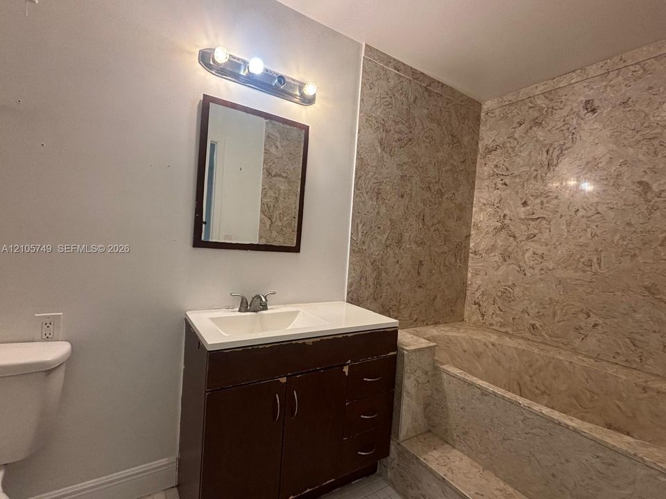 Bathroom featuring a vanity with an integrated sink, three-light wall-mounted fixture, framed mirror, and a tub enclosure with stone-patterned surround
