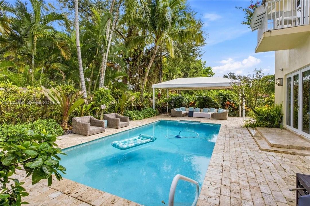 In-ground swimming pool with stone paver decking and a covered outdoor structure, bordered by established tropical landscaping