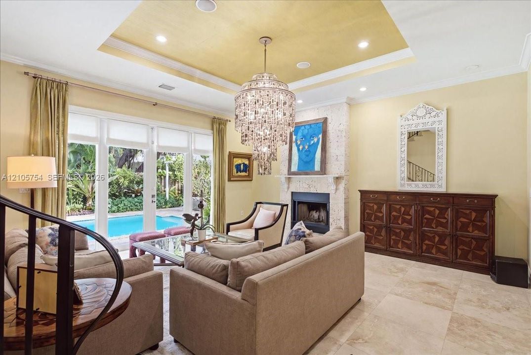 Tray ceiling with recessed lighting, oversized crystal chandelier, floor-to-ceiling glass panel doors, stone-faced fireplace with hearth, and large-format tile flooring