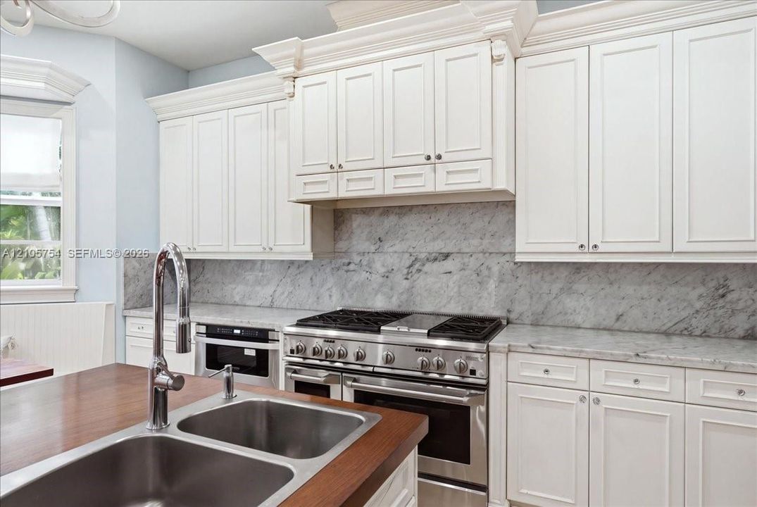 Custom white cabinetry with crown molding, natural stone backsplash and countertops, professional-grade stainless steel gas range, and wood-topped island with an integrated stainless steel dual-basin sink