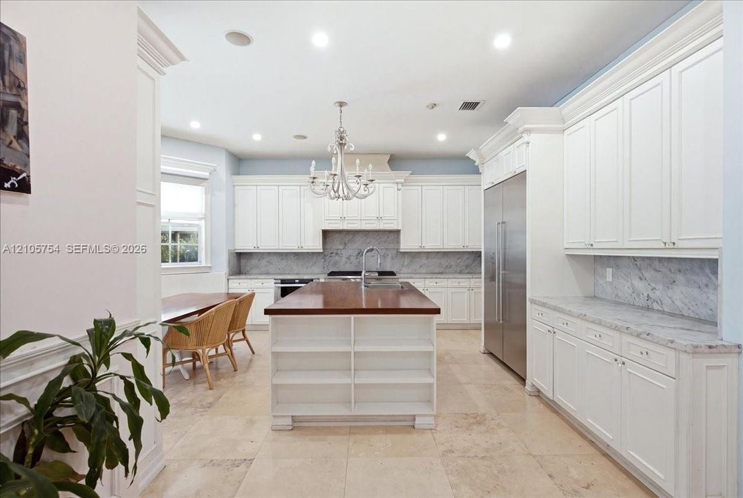 Kitchen featuring custom white cabinetry, central island with wood countertop, stainless steel refrigerator, stone tile flooring, and stone backsplash