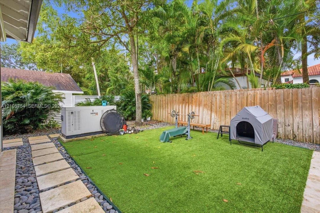 Backyard featuring artificial turf, natural stone paver pathway, decorative pebble border, wooden privacy fencing, and a permanent exterior generator