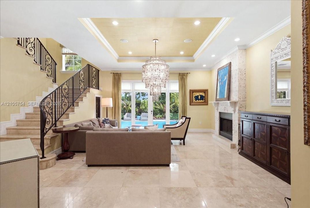 Living area featuring large-format tile flooring, a recessed ceiling with a central crystal chandelier, a fireplace with textured stone surround, and a wrought-iron staircase railing