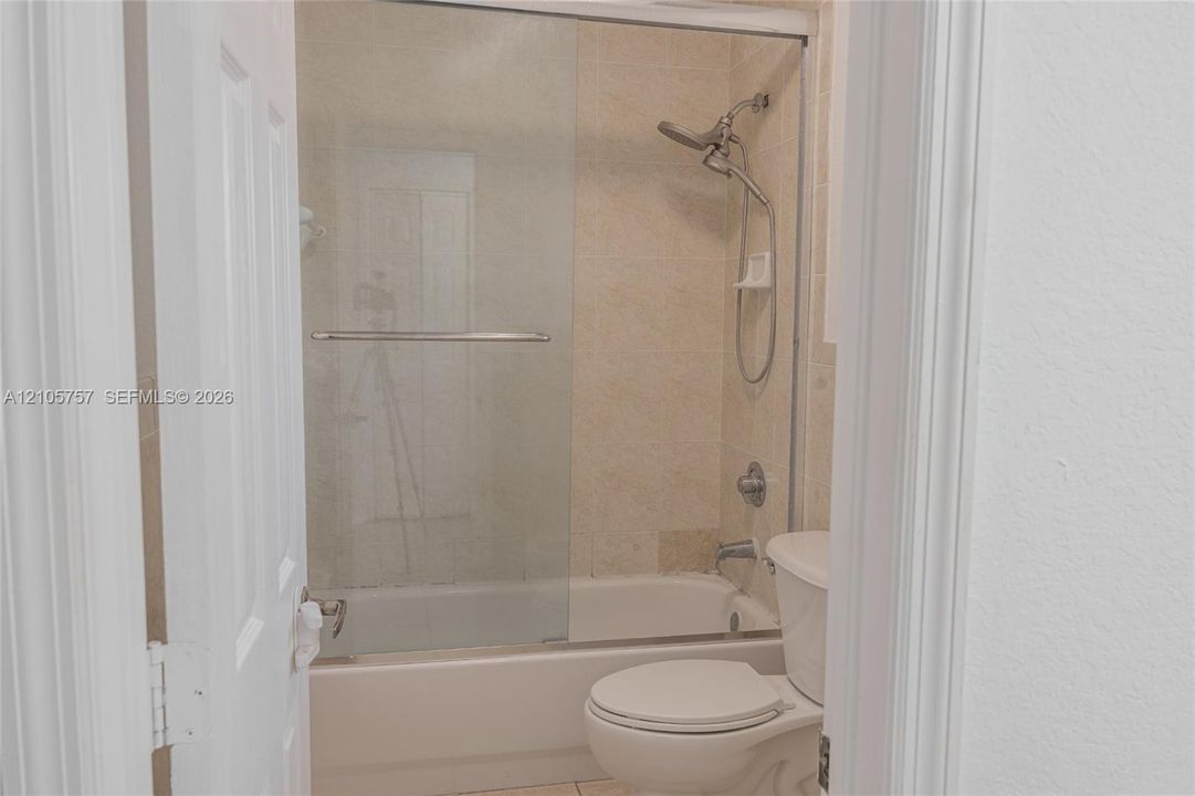 Bathroom featuring a tiled tub-shower combination, glass sliding enclosure, adjustable handheld shower fixture, and a white porcelain commode