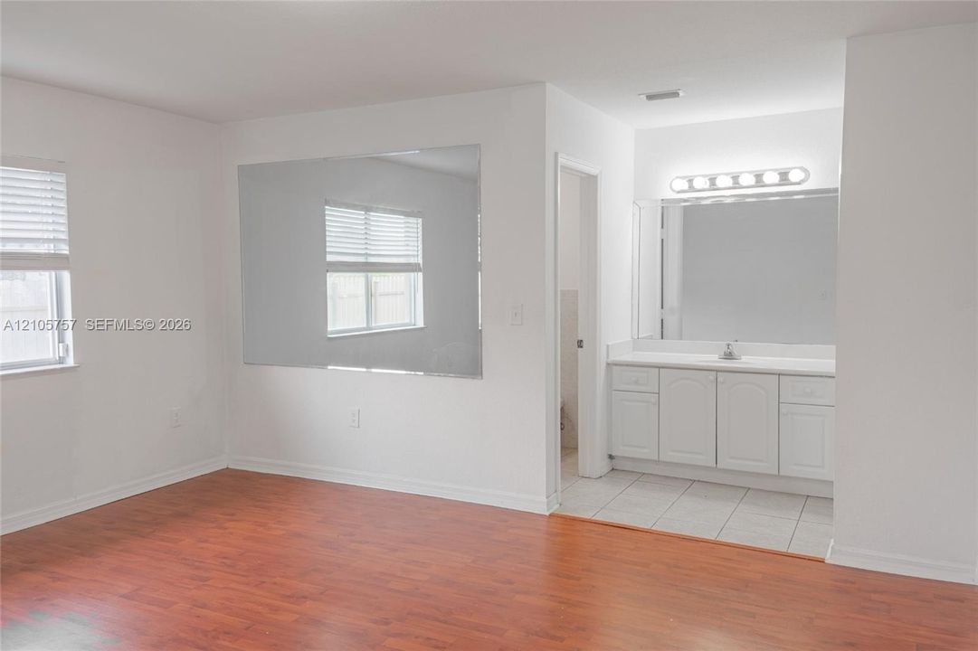 Wood-finish flooring, large wall-mounted mirror, white vanity cabinetry with surface-mount countertop, and horizontal slat window blinds