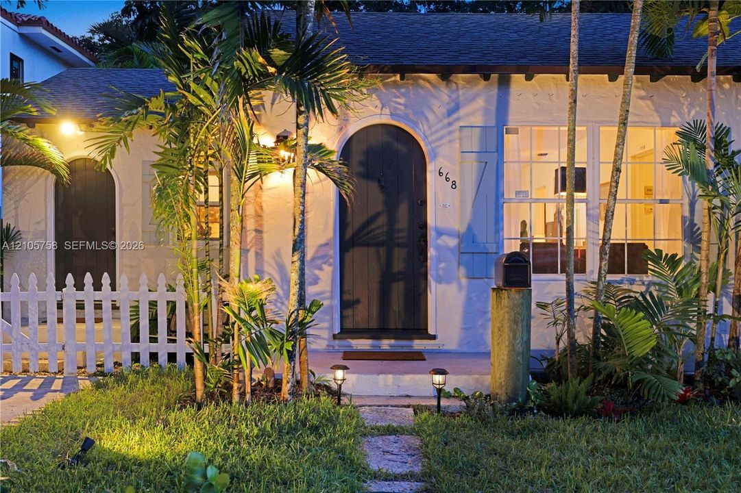 Stucco exterior featuring an arched entryway, shuttered multi-pane windows, a stone walkway, and a white picket fence