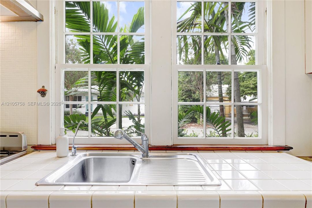 Double-basin stainless steel sink with a high-arch pull-down faucet, white tile countertop, and multi-pane double-hung windows