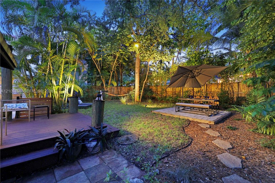 Backyard featuring a raised wood-finish deck, paved patio area, stone paver path, mature trees, and privacy fencing