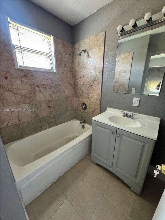 Bathroom featuring a tub with stone-tiled surround, vanity with an integrated sink, wall-mounted mirror with multi-bulb light fixture, and tile flooring
