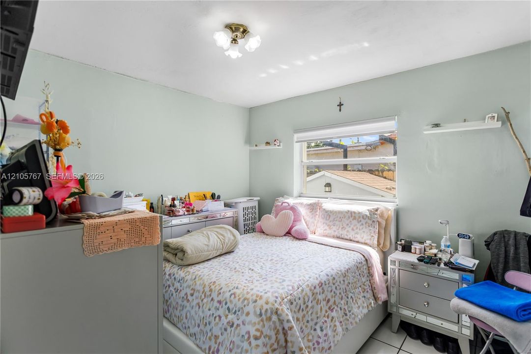 Bedroom featuring light-toned walls, tile flooring, a multi-light flush-mount ceiling fixture, a window with an integrated roller shade, and wall-mounted shelving
