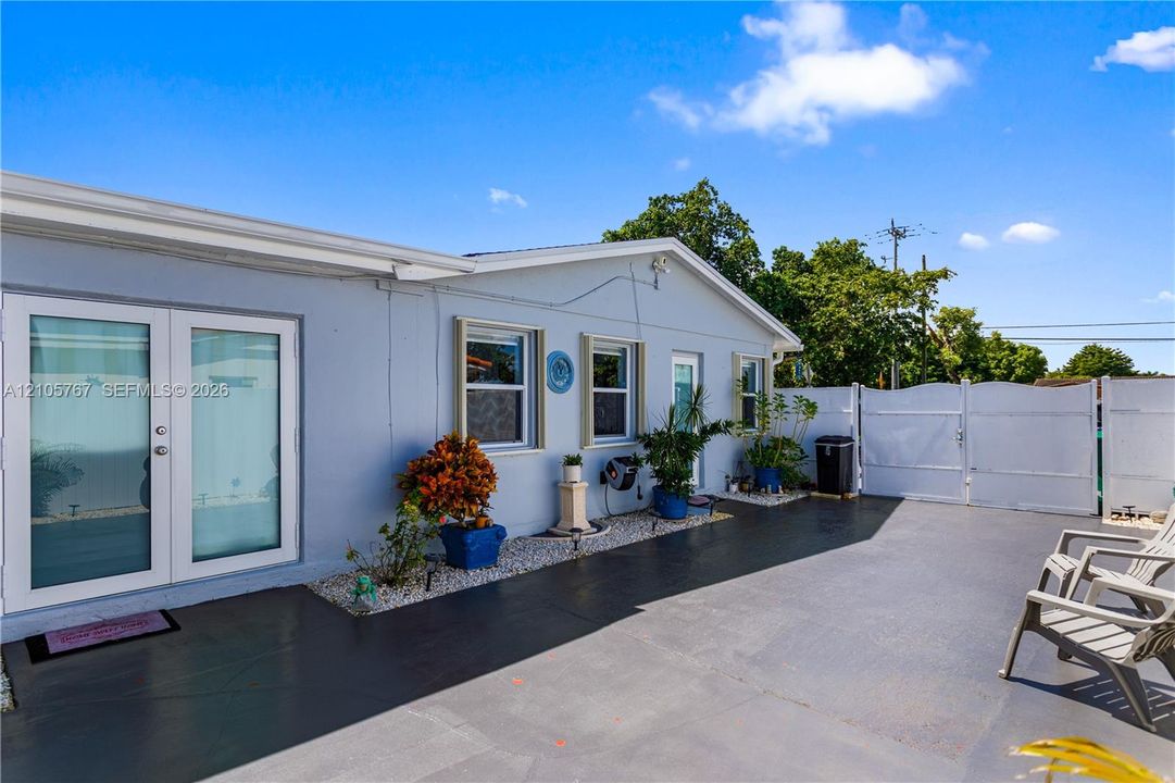 Exterior wall with light blue finish, dual glass-paneled exterior doors, light-colored framed windows, paved patio surface, and a perimeter fence with a gated entry